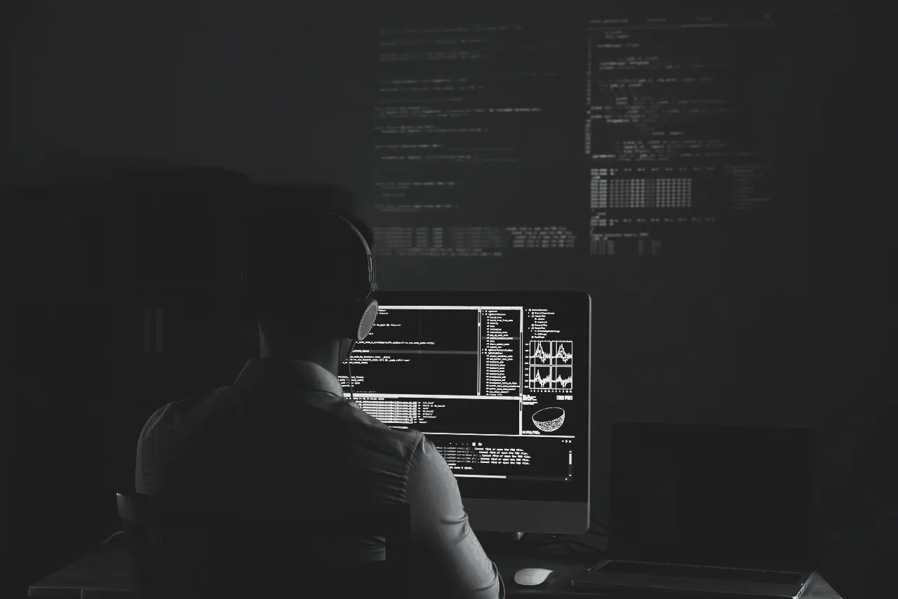 Person vor mehreren Bildschirmen mit Programmcode in einem dunklen Raum